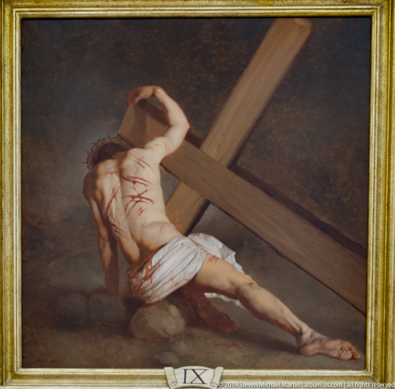 Framed devotional painting of wounded Christ bent beneath a large wooden cross, with a small IX label below.