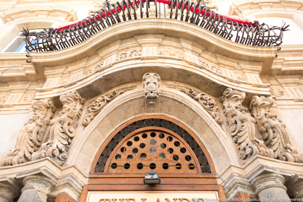Upward view of an ornate pale-stone portal with carved scrolls, masks, curved pediment, and iron balcony above.