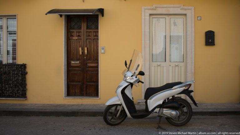 White motor scooter parked against an ochre wall between a dark wooden door and a narrow pale-framed doorway.
