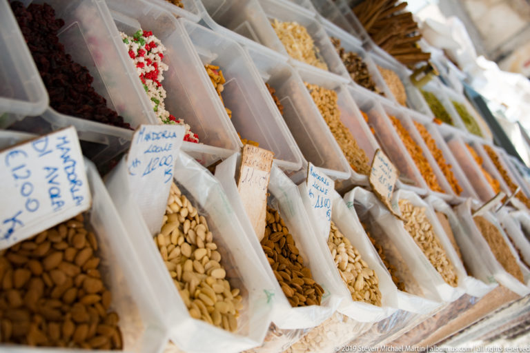 Rows of clear market bins filled with dried legumes, nuts, and seeds, with handwritten Italian price labels.