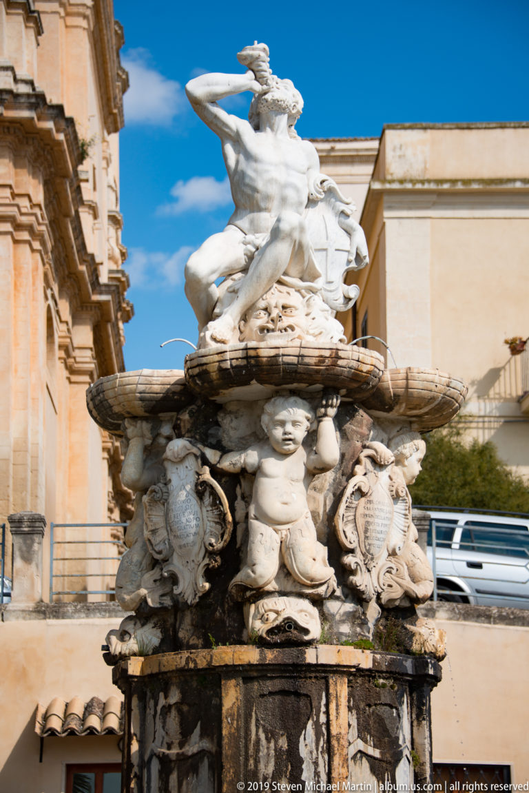 Multi-tier stone fountain with winged figure and sea creature above putti, shields, and water-stained pedestal.