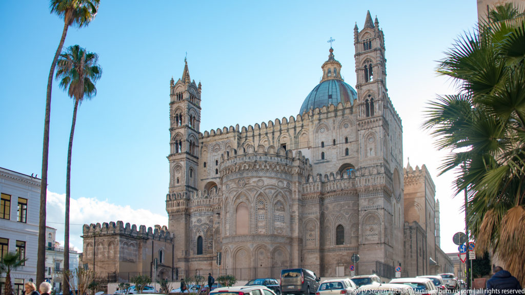 Wide street-level view of Palermo Cathedral showing the patterned apse, dome, towers, palms, vehicles, and pedestrians.