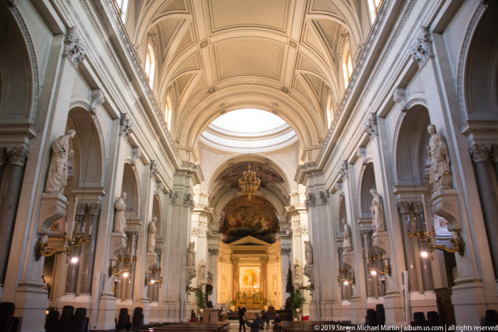 Closer axial view along the cathedral nave toward the bright crossing, sanctuary, statues, and painted apse.