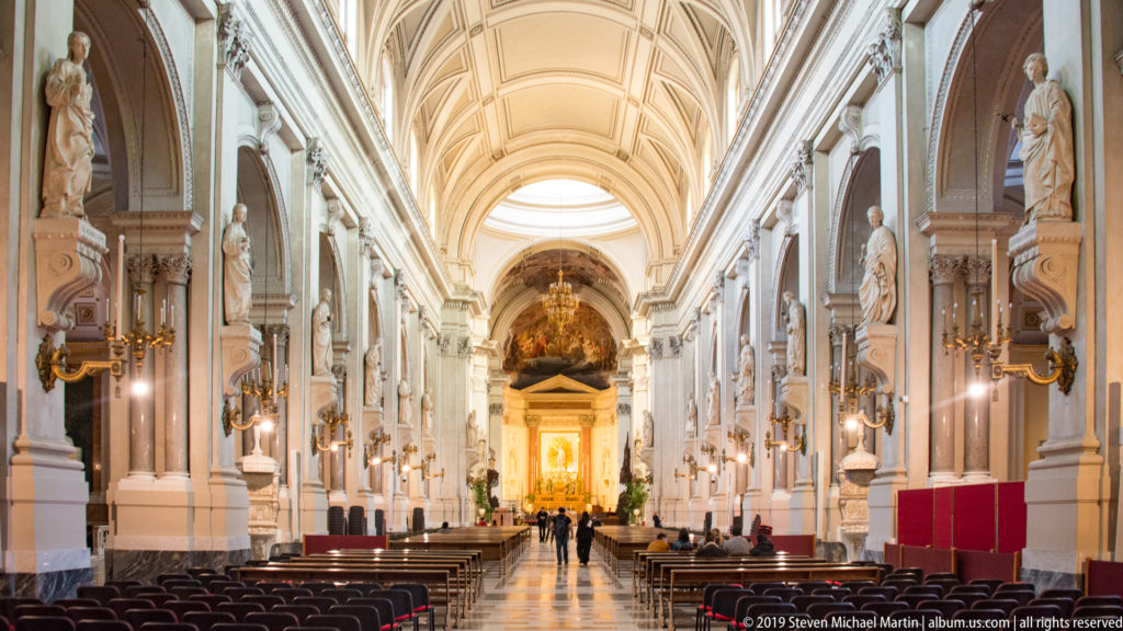 Visitors walking through the long cathedral nave toward a bright sanctuary beneath a pale vaulted ceiling.
