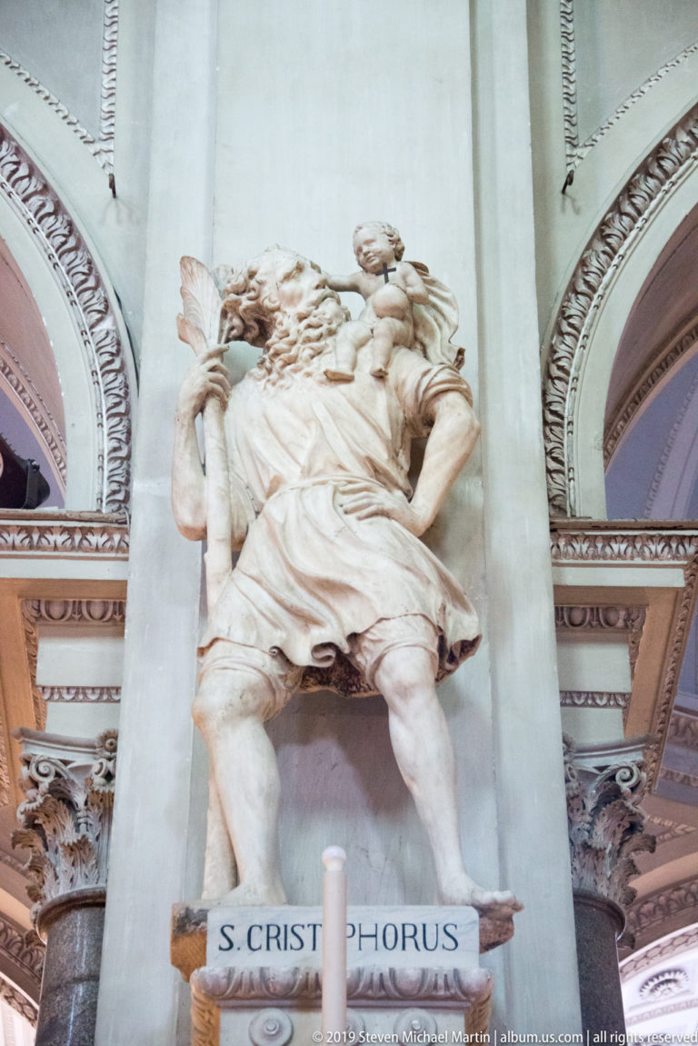 Large pale stone statue of Saint Christopher carrying the Christ Child, mounted on a cathedral pier above an inscribed base.