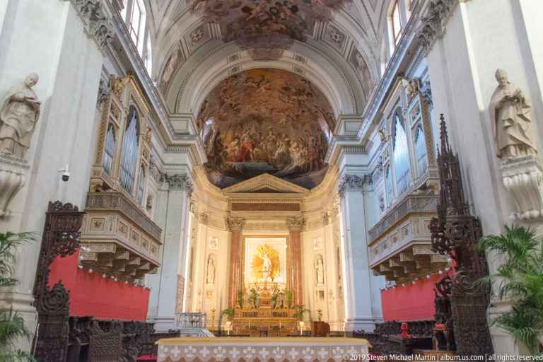 Cathedral sanctuary with altar, bright devotional image, dark carved furnishings, statues, and a painted apse above.