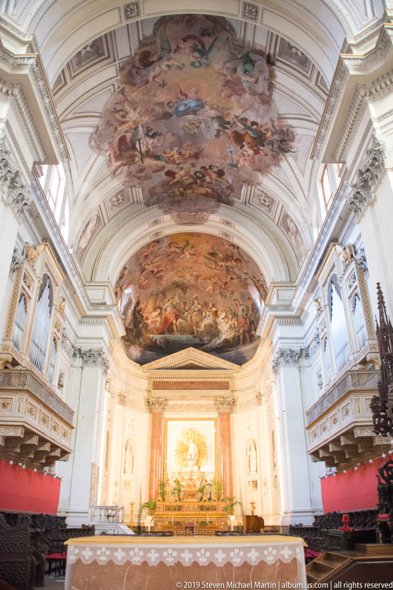 Centered sanctuary view with painted vaults overhead, organ lofts on both sides, red coverings, and illuminated altar beyond.