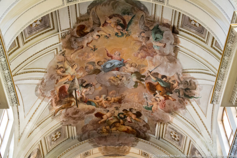Large painted vault scene with airborne figures and clouds in warm tones, framed by pale ribs and decorative panels.