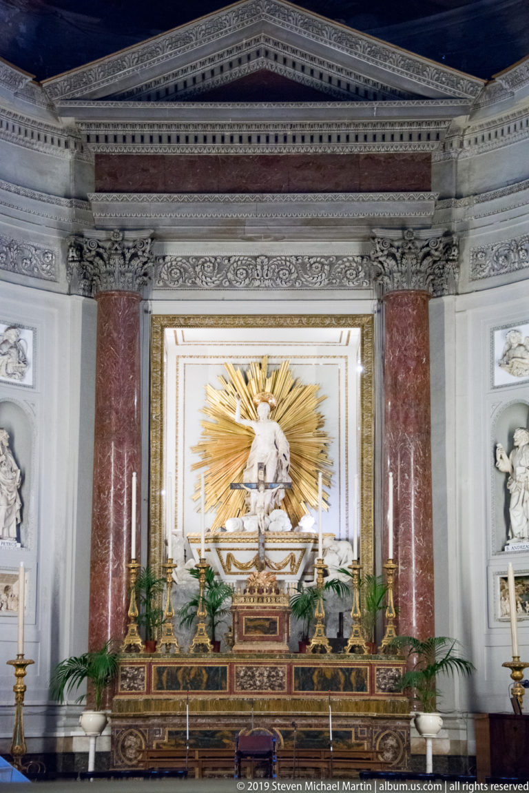 Side altar framed by red marble columns, with gold sunburst devotional image, candlesticks, plants, and flanking niches.