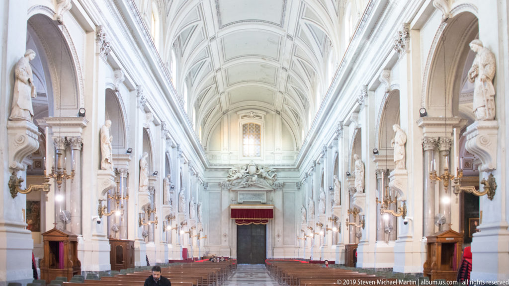 Long symmetrical nave with pale coffered vault, repeated arches and statues, wooden pews, and a visitor near center.