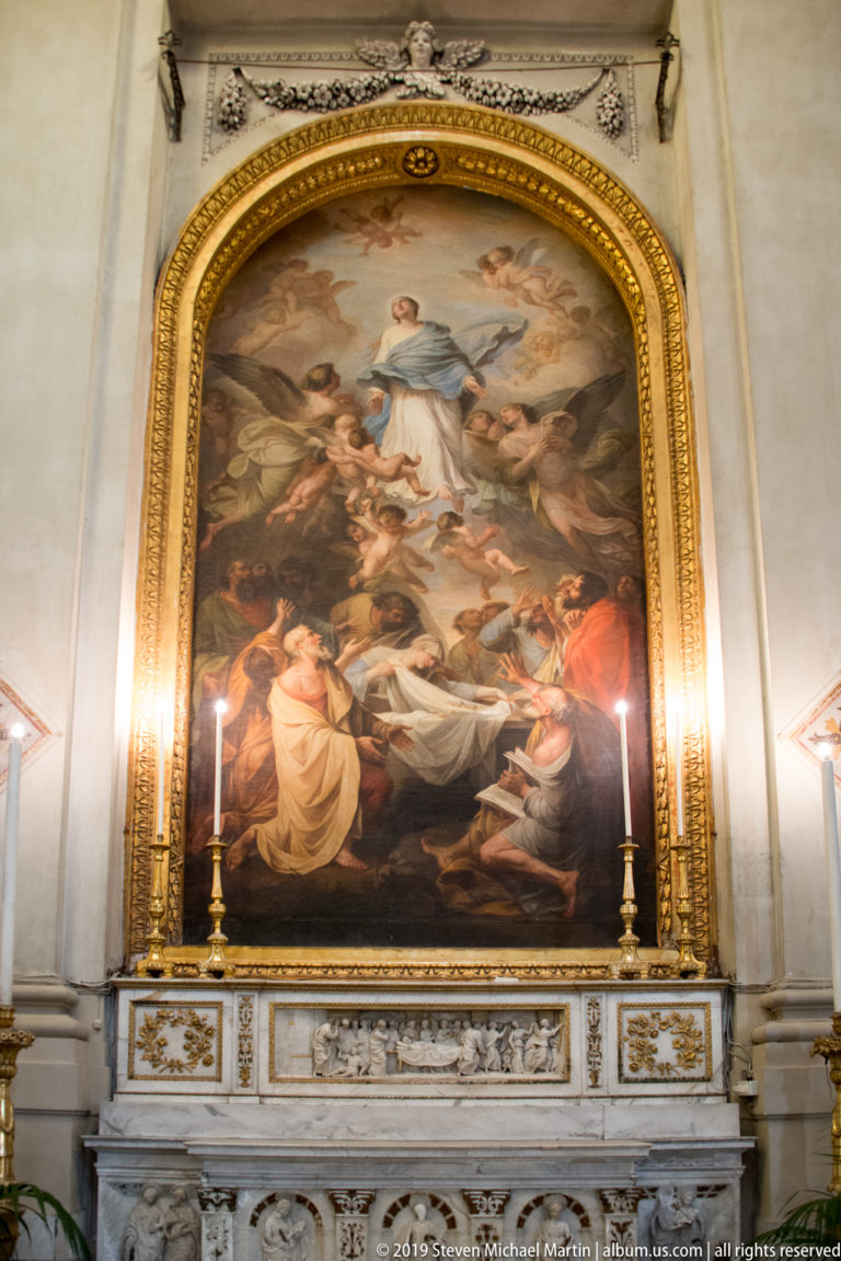 Large arched religious painting in a gilded frame above a marble altar, flanked by tall candles.