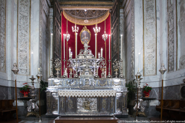 Elaborate silver-toned chapel altar with central oval devotional object, red backdrop, candlesticks, lamps, and marble walls.