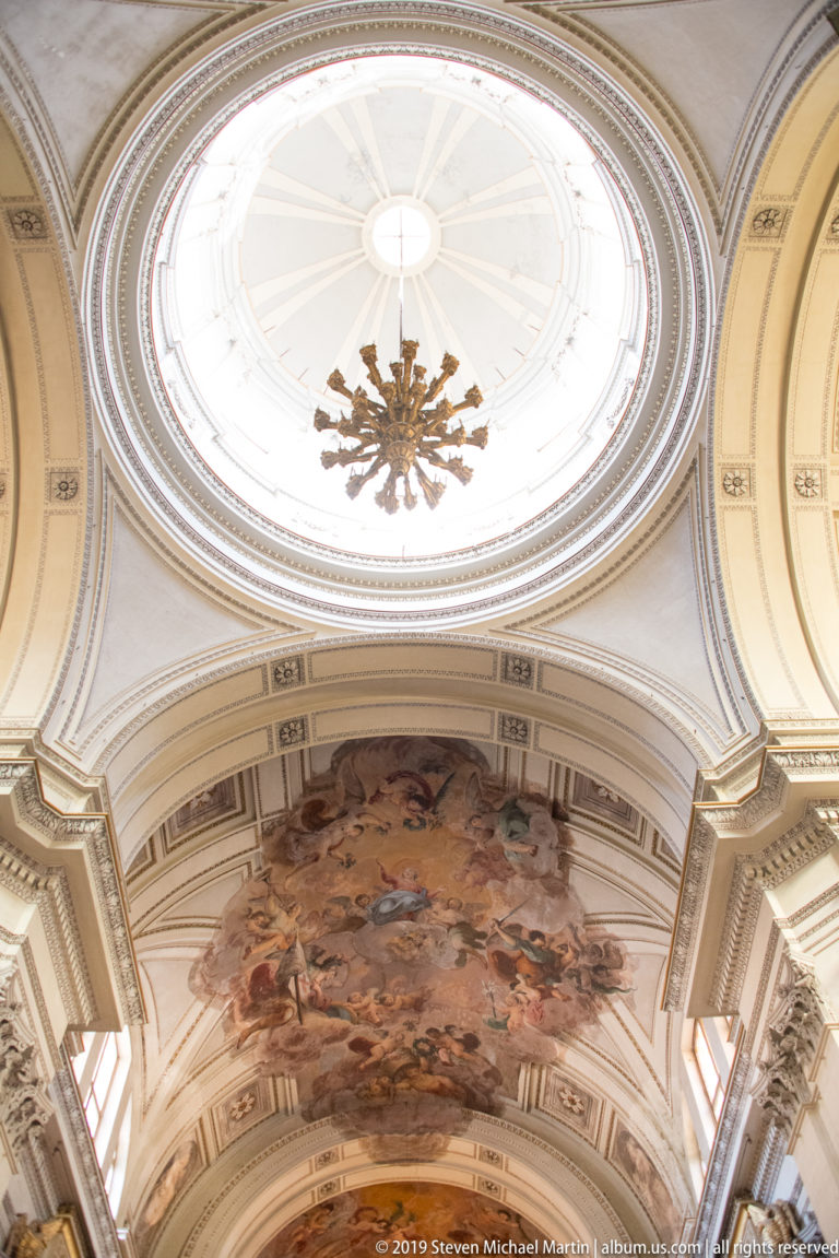 Upward view of an oval skylight above a large painted vault framed by pale moldings and gilded decoration.