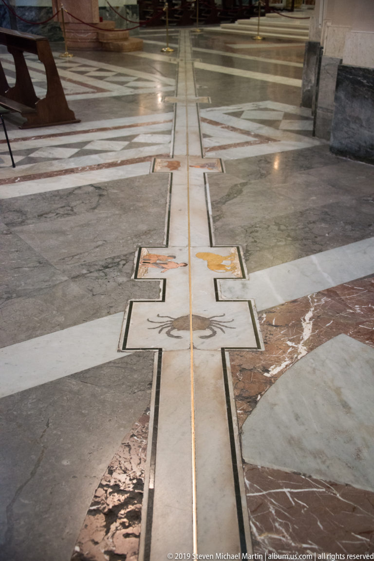 Geometric cathedral pavement of white, gray, red, and dark marble bands crossing polished stone panels.