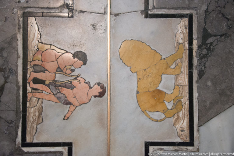Close view of two colored figurative stone inlays set into a pale marble floor panel with visible wear.