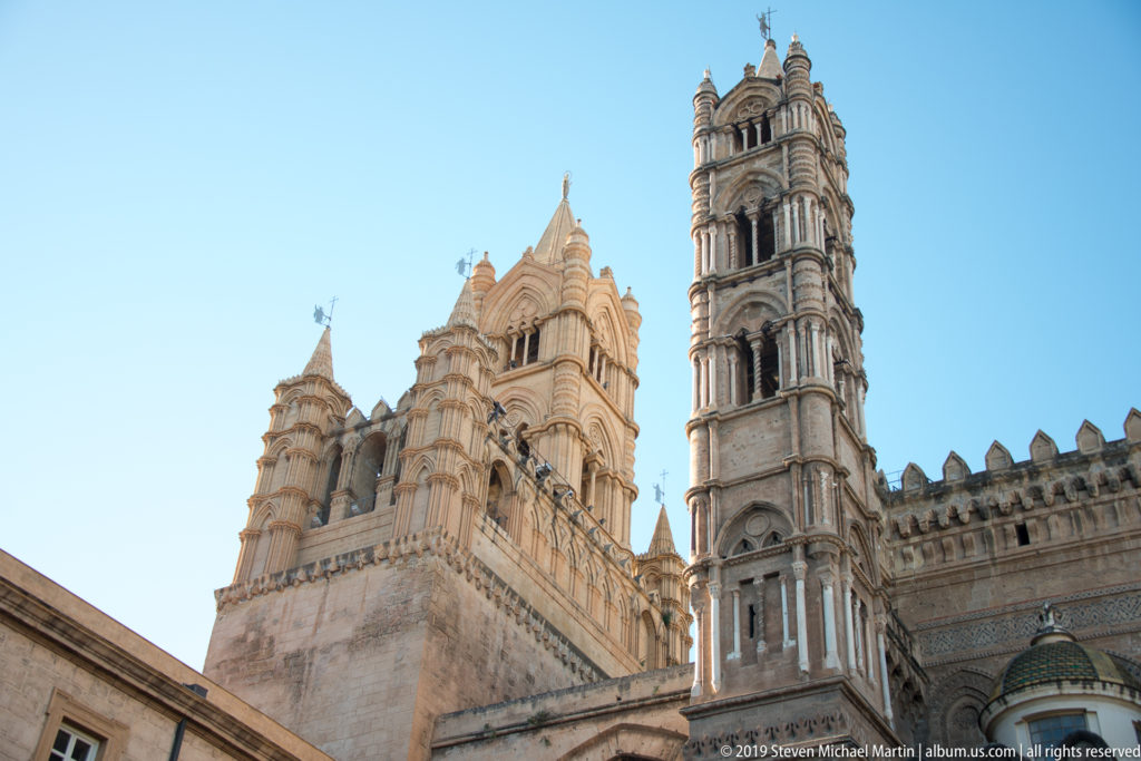 Low-angle view of Palermo Cathedral’s ornate multi-tiered towers and crenellated wall against blue sky.