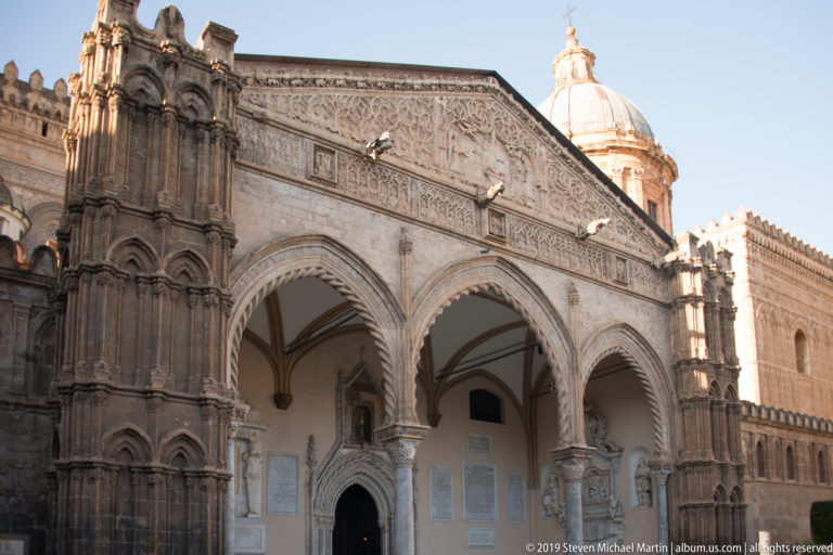 Three richly carved pointed arches of Palermo Cathedral’s portico, with sculpted frieze above and dome behind.