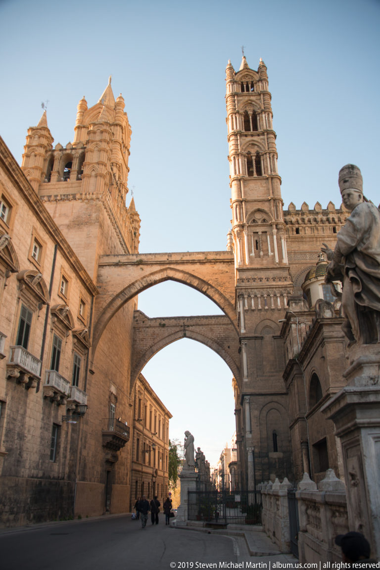 Tall cathedral bell tower beside a street, connected across the roadway by two broad masonry arches.