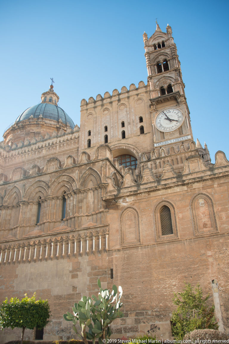 Broad exterior view of Palermo Cathedral with dome, crenellated side wall, clock tower, blind arches, and garden plants.