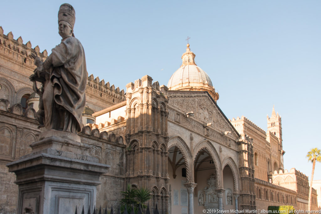 Oblique view of Palermo Cathedral’s three-arched portico, dome, crenellated walls, towers, and foreground statue.