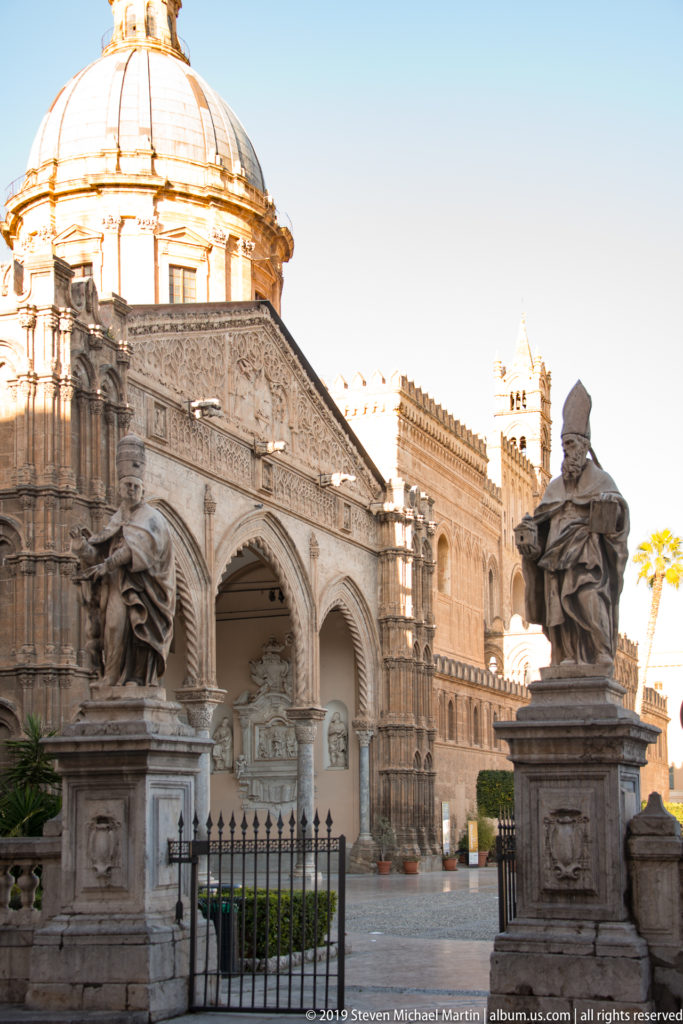 Arcaded cathedral portico with dome behind, flanked by pedestal statues, iron gate, and forecourt garden.