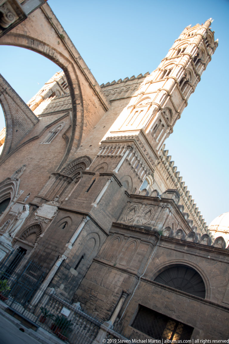 Steep low-angle view up Palermo Cathedral’s masonry façade toward an ornate tower against blue sky.