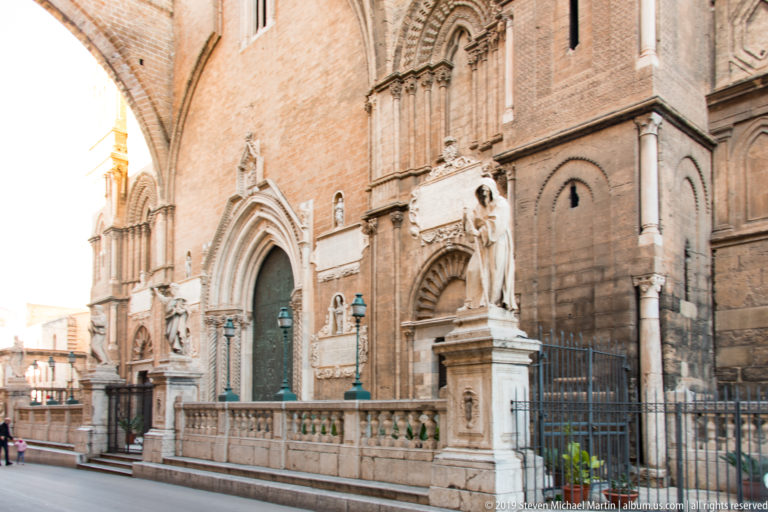 Oblique view along Palermo Cathedral’s side façade with pointed portal, statues, iron railings, and warm masonry.