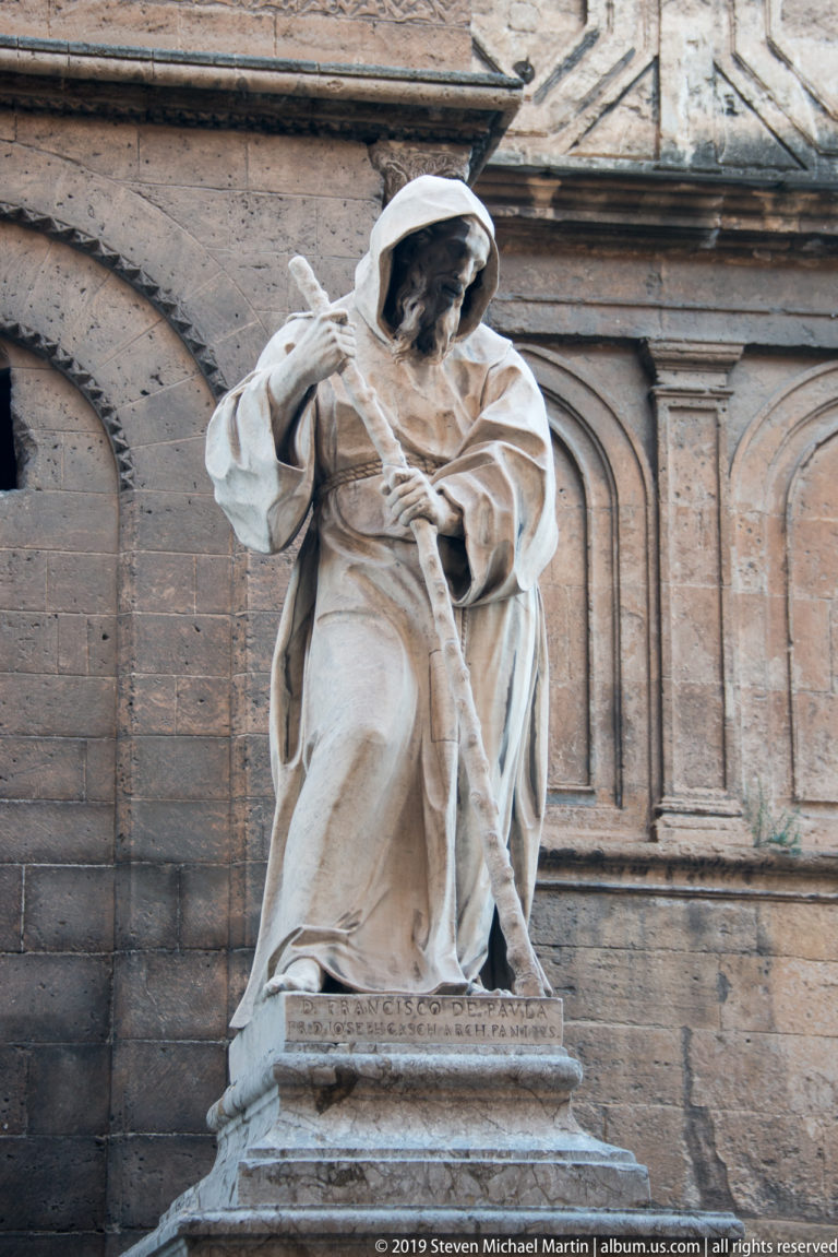 Hooded marble female religious figure with downcast head and folded robe against a rusticated cathedral wall.