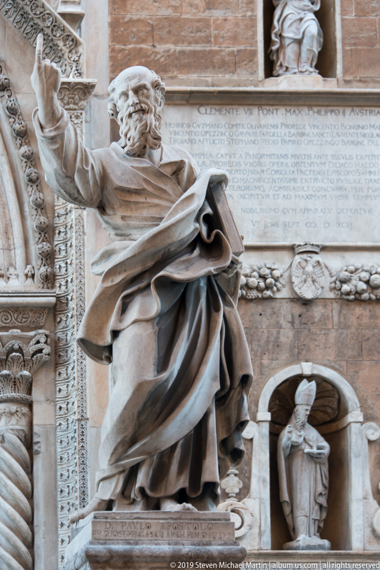 Bearded marble apostle extending one arm beside an inscription, carved portal edge, and small niche statue.