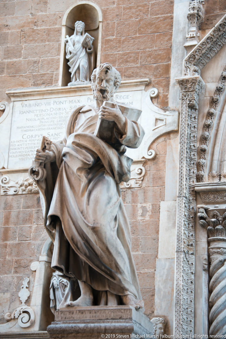 Robed marble apostle holding an attribute before a large inscribed wall panel, with a small niche statue above.