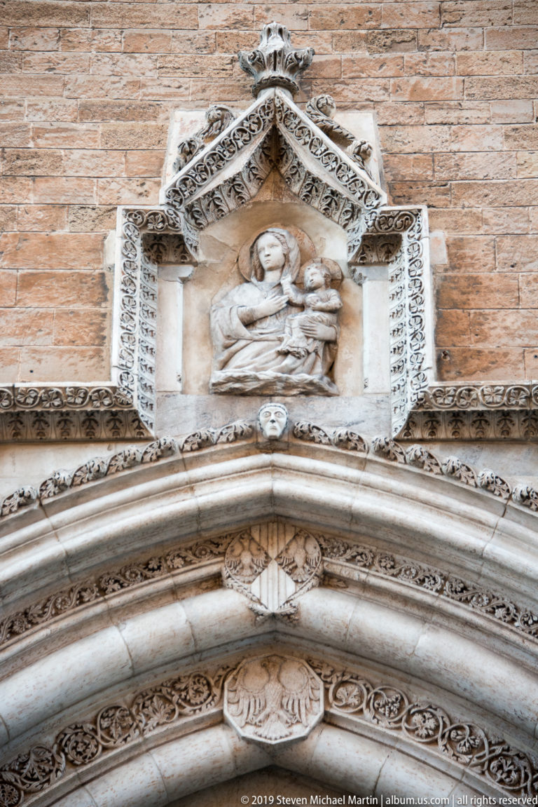 Small sacred relief in an ornate gabled frame above the carved pointed arches of a cathedral portal.