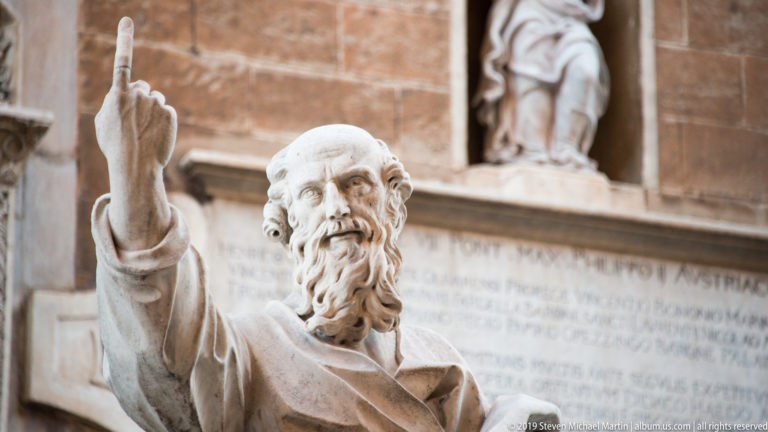 Close view of a bearded marble apostle raising one hand, with inscription and niche sculpture behind him.
