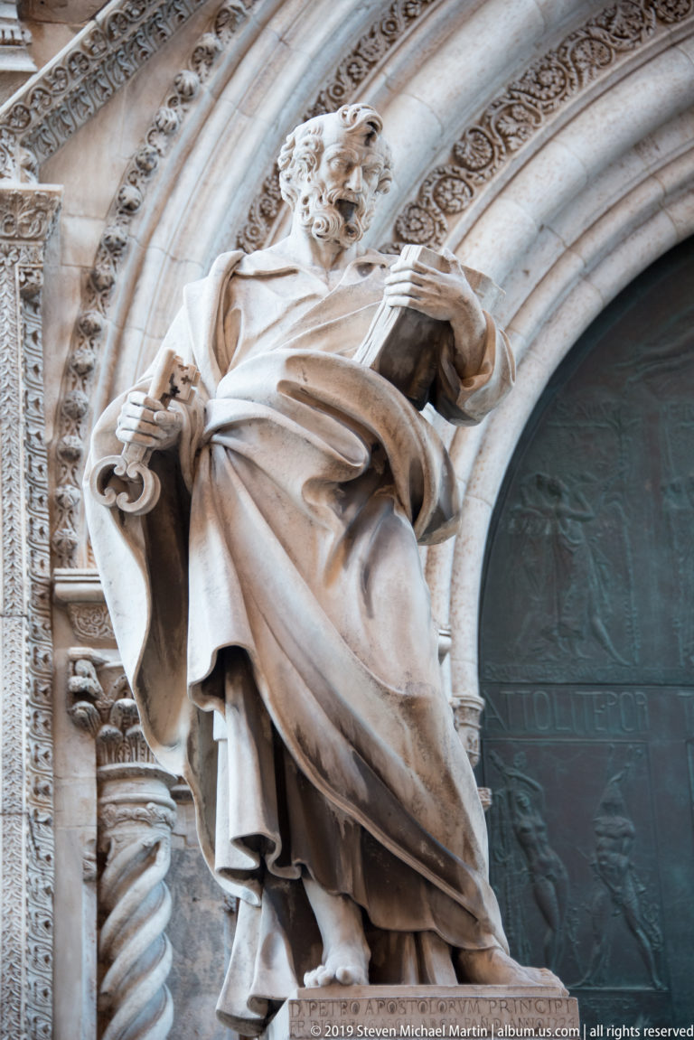 Low-angle view of a robed marble apostle statue beside the cathedral’s carved portal and patterned archivolts.