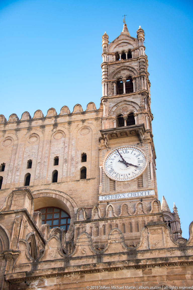 Upward view of Palermo Cathedral exterior with large round clock, open-arched tower, crenellations, and carved stonework.