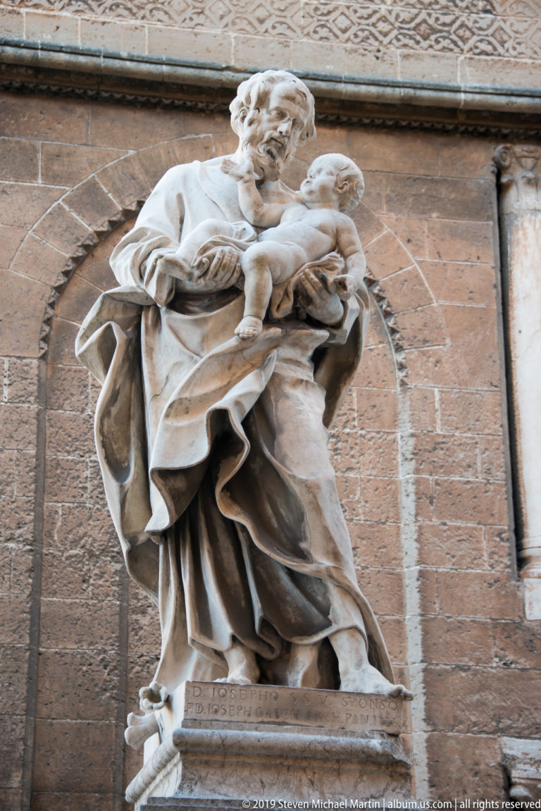Marble religious figure holding a child, with deeply carved drapery, standing against the cathedral’s warm stone wall.