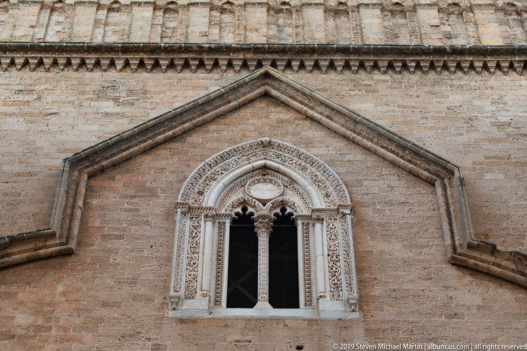 Paired arched cathedral window beneath a pointed gable, surrounded by intricate pale stone carving on warm masonry.