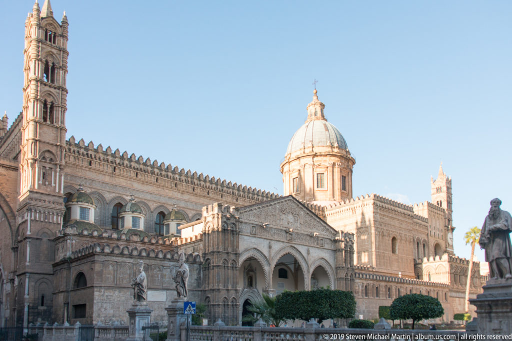 Oblique sunlit view of Palermo Cathedral with arcaded entrance, dome, towers, crenellated roofline, and foreground statue.