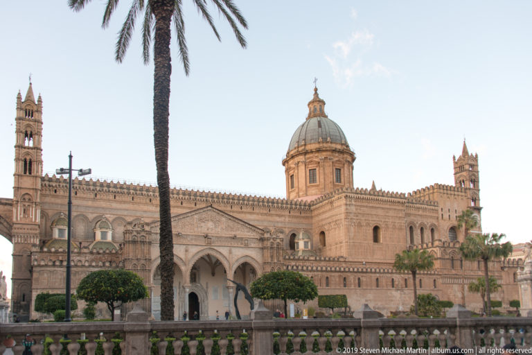 Broad exterior view of Palermo Cathedral with dome, arcades, towers, crenellations, palm tree, and landscaped forecourt.