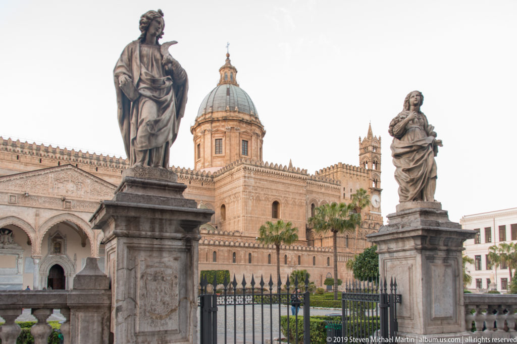 Two stone statues on pedestals frame Palermo Cathedral’s dome, arcades, iron fence, and landscaped forecourt.