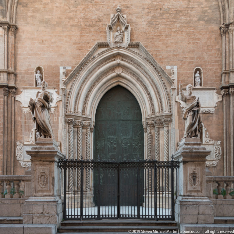 Pointed cathedral portal with carved archivolts, bronze door, iron gate, and marble saint statues on pedestals.