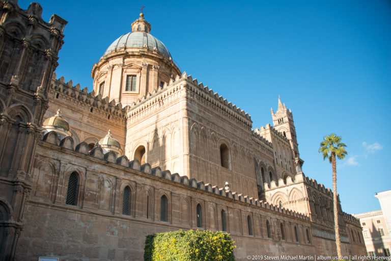 Low-angle exterior view of Palermo Cathedral’s long stone side elevation, crenellations, dome, and tower details under blue sky.