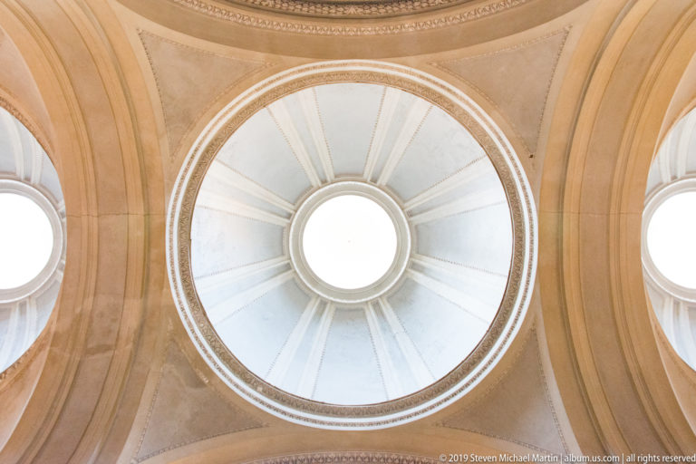 Upward view into a pale dome with circular central oculus, radial ribs, concentric molding, and side windows.