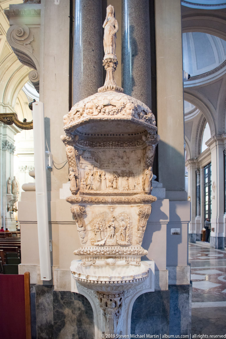 Ornately carved pale stone font with relief decoration, domed cover, and small crowning figure inside the cathedral.