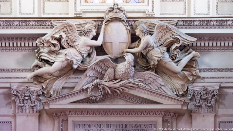 Ornate pale marble wall monument with winged figures, eagle, central tablet, pediment, and Latin inscription below.