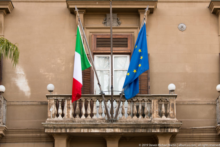 Italian and European Union flags hang from poles above a stone balcony and shuttered window on a warm-toned palace façade.