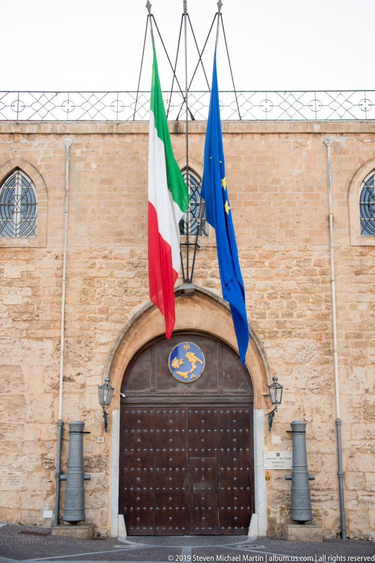 Large dark wooden arched gate beneath Italian and European Union flags, with a small blue-and-gold emblem above.