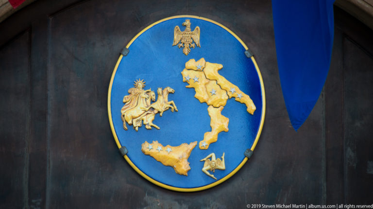 Blue oval plaque with raised gold map of Italy and Sicily, small gilded symbols, and an eagle above.