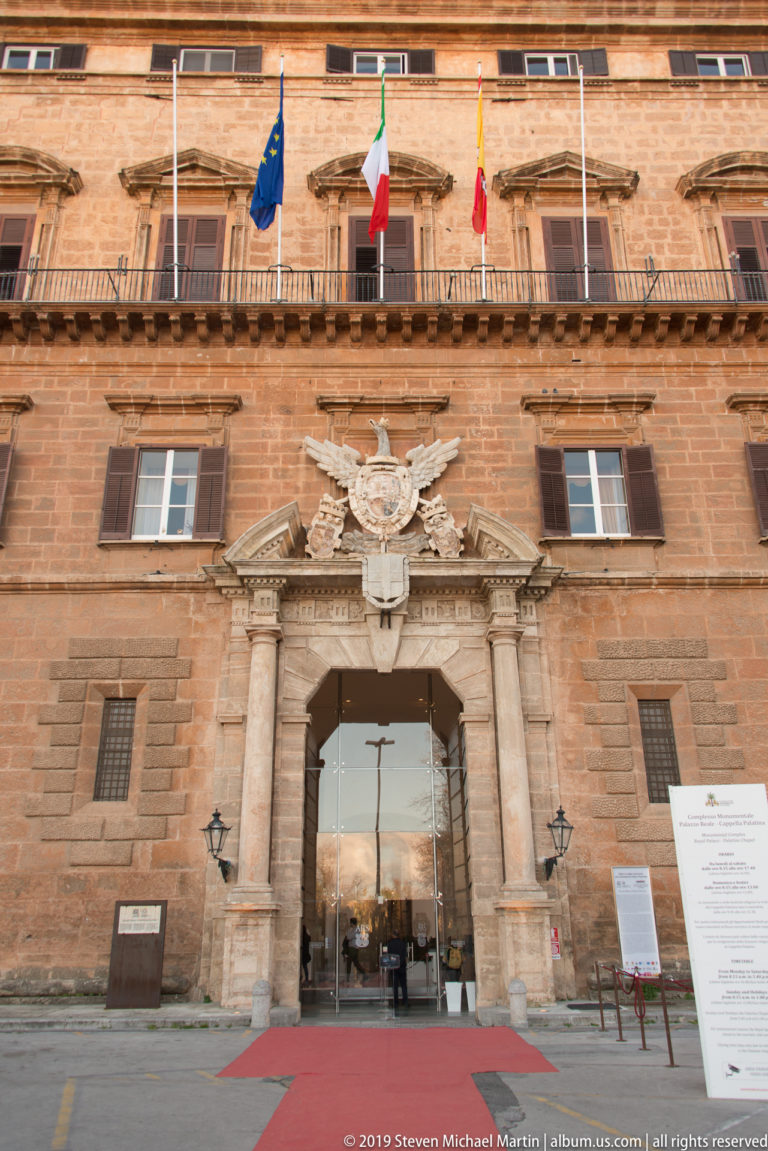 Frontal palace entrance with paired columns, heraldic relief, balcony flags, red carpet, and rusticated reddish stone façade.