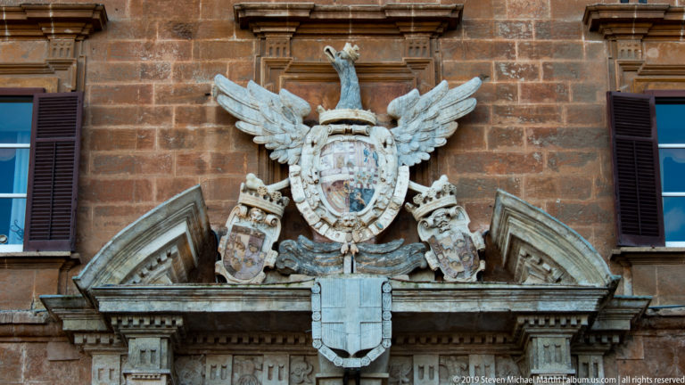 Large carved coat of arms with crowned shield and spread wings mounted on the reddish stone façade between shuttered windows.