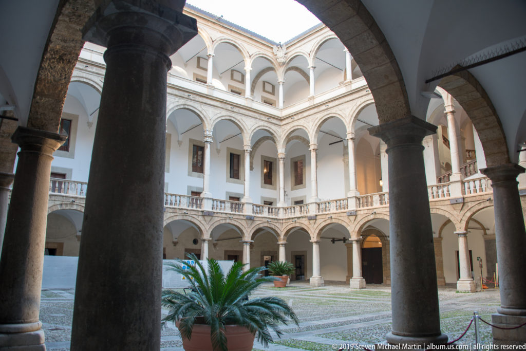 Large dark columns frame a bright two-level arcaded courtyard with patterned paving and a potted cycad.